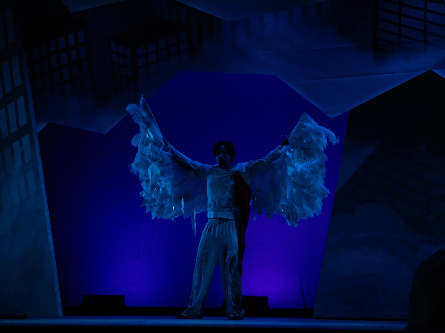 A performer spreads large feathered wings on the blue-lit Giants Have Us in Their Books stage